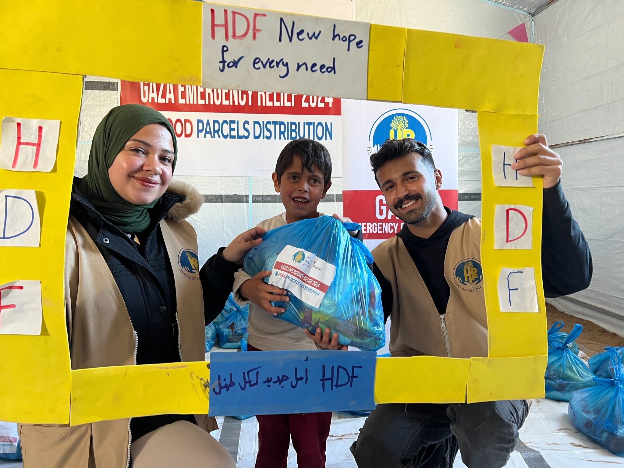 A child holding an HDF food parcel between two team members at a Gaza food distribution in 2024