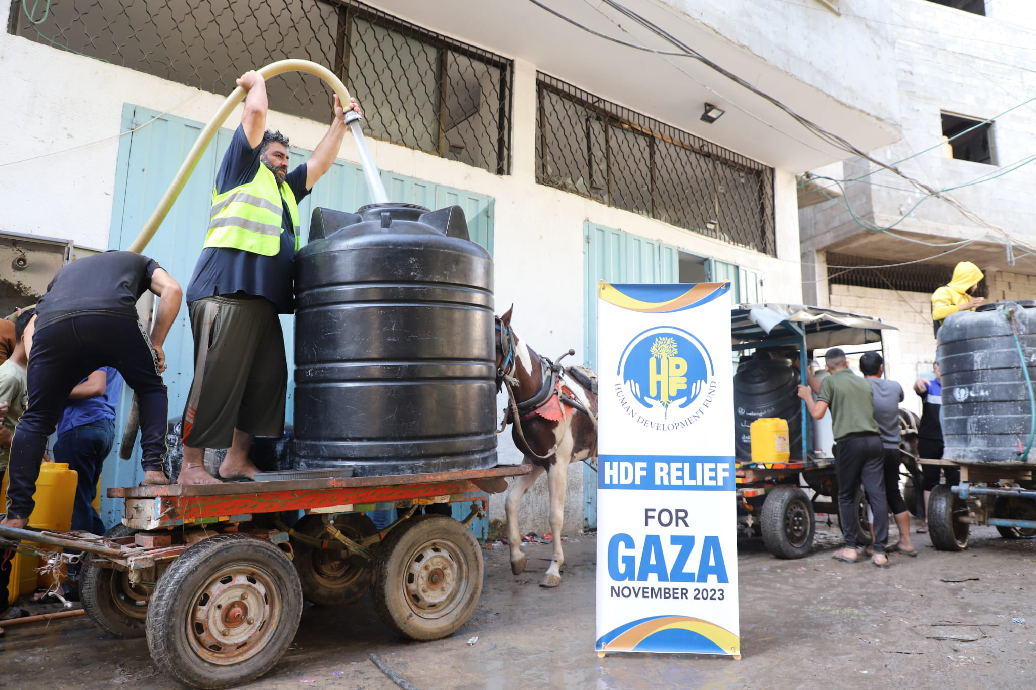 A worker fills a water tank on a cart beside an HDF Relief for Gaza banner dated November 2023