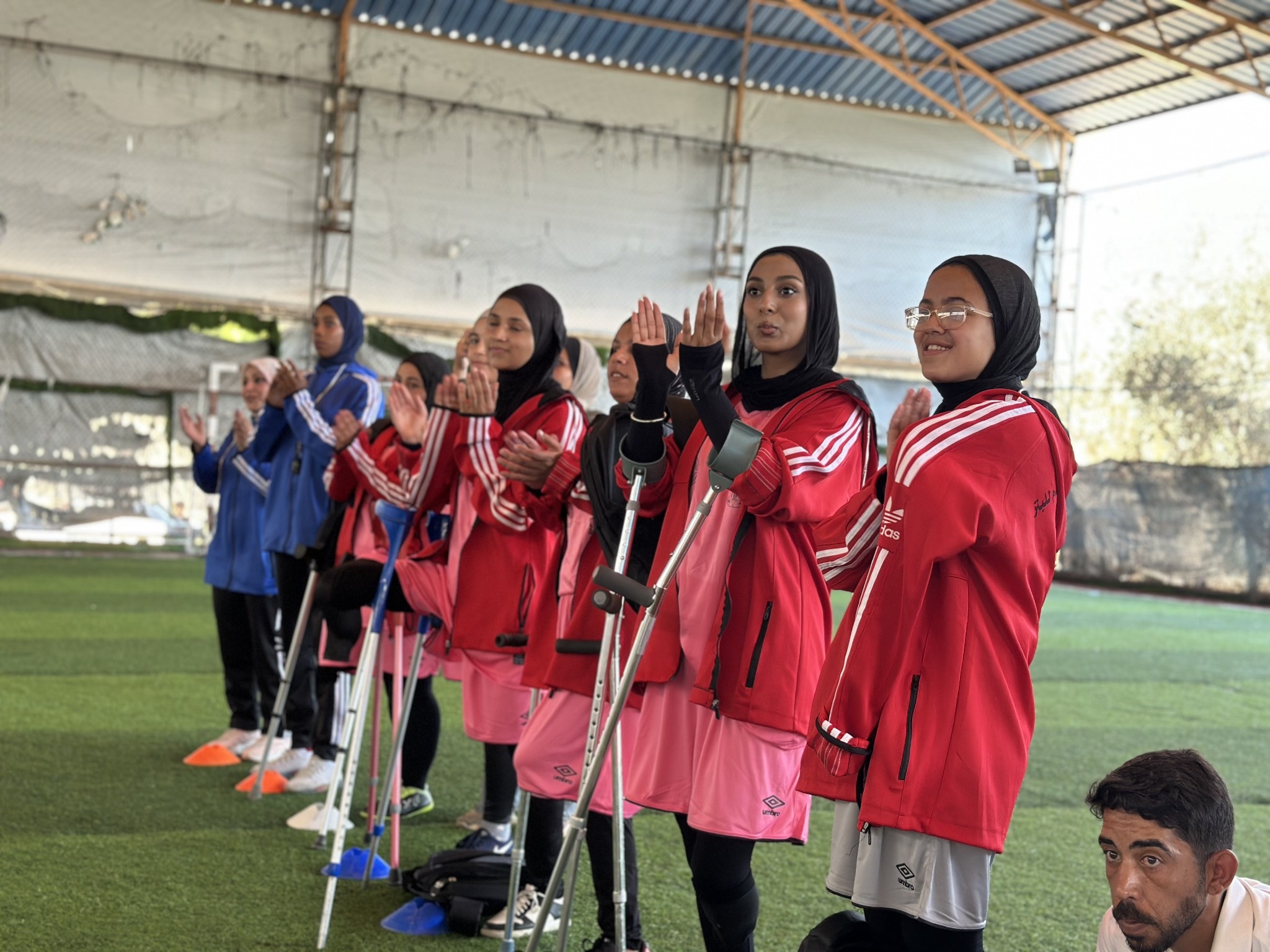 Women football players in red and blue tracksuits applaud on a covered pitch, several using forearm crutches