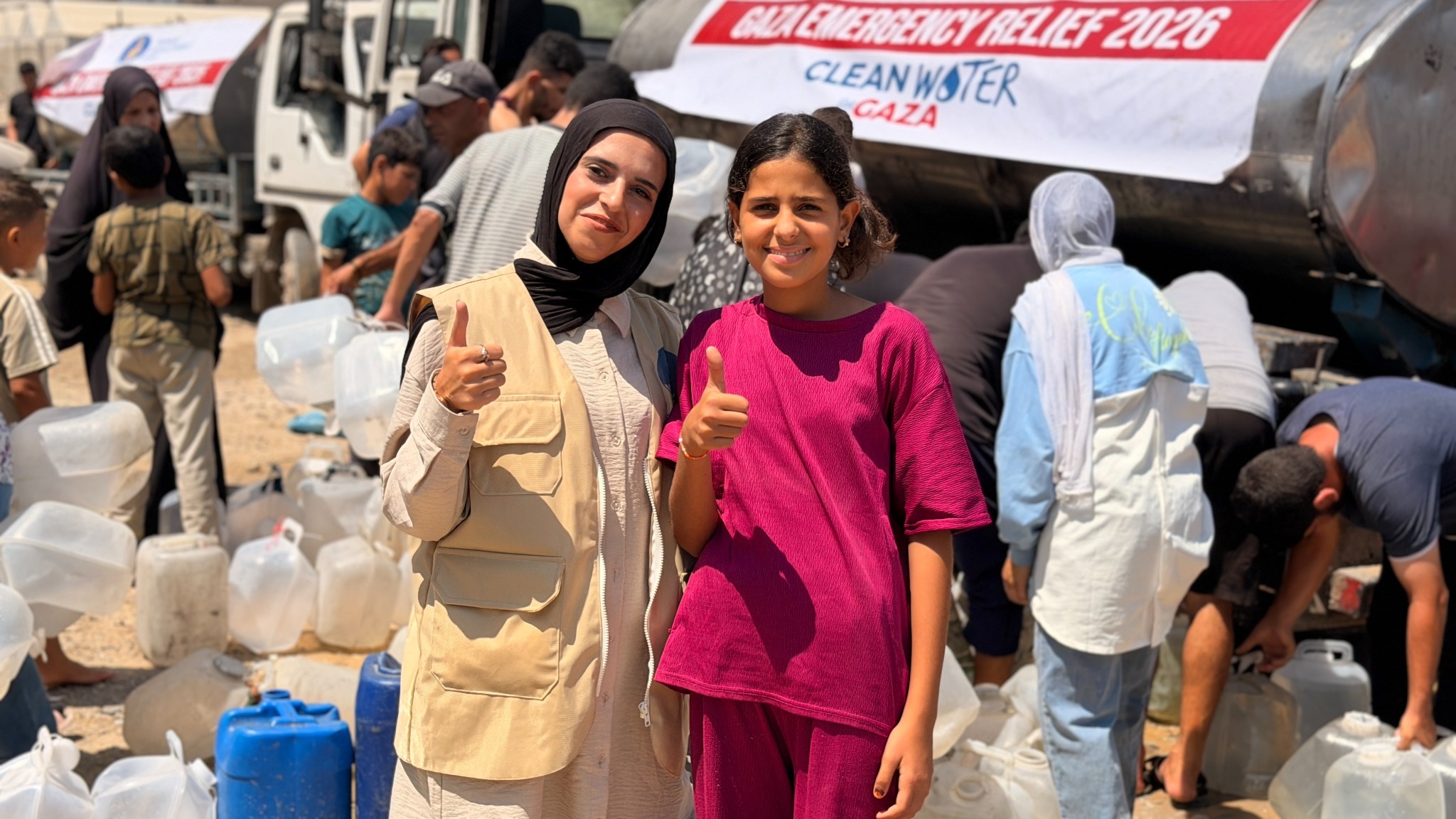 A woman and girl give thumbs-up beside a water tanker and rows of containers.