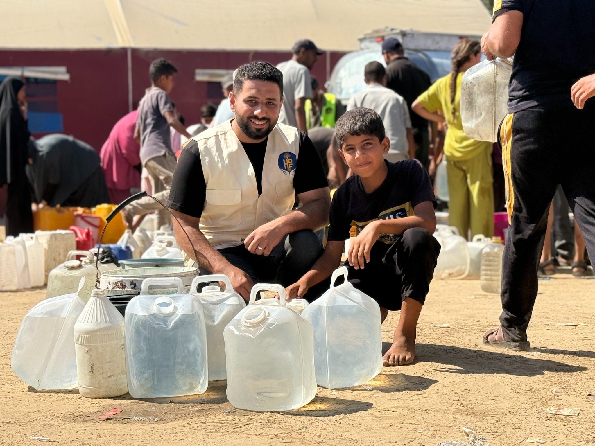 A man and boy crouch beside water containers while people gather behind them.