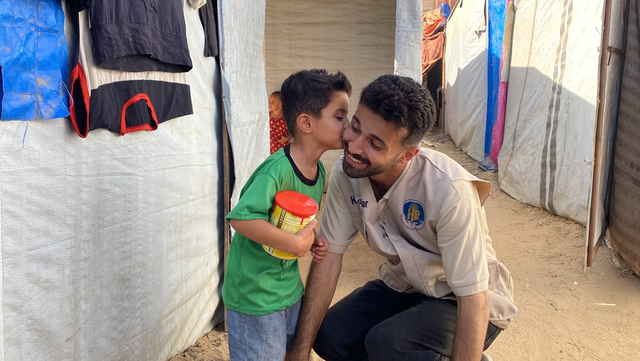 A child holding a red-lidded container kisses a smiling man’s cheek between tarp shelters.