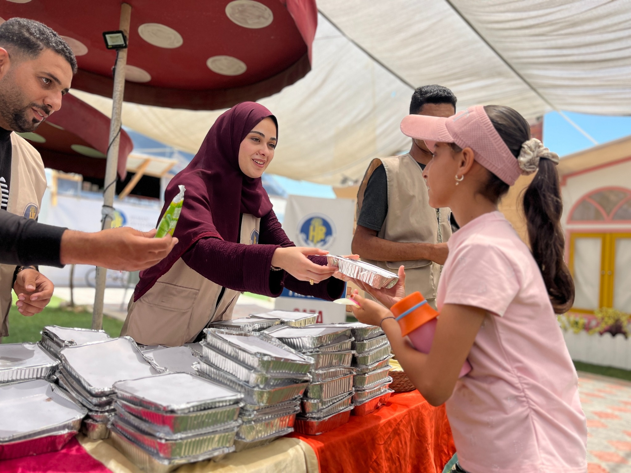 A woman hands a foil meal tray to a girl across a table stacked with meals.