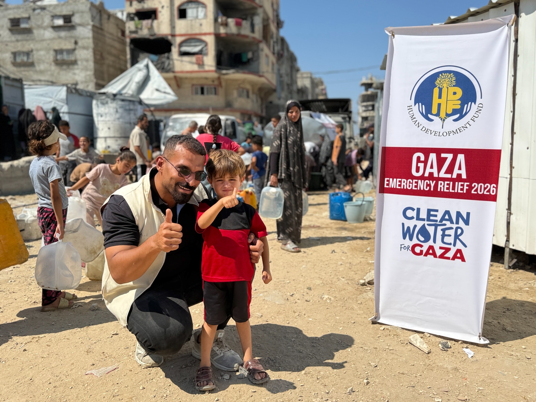 A man and boy pose beside a clean-water relief banner as people carry containers behind them.