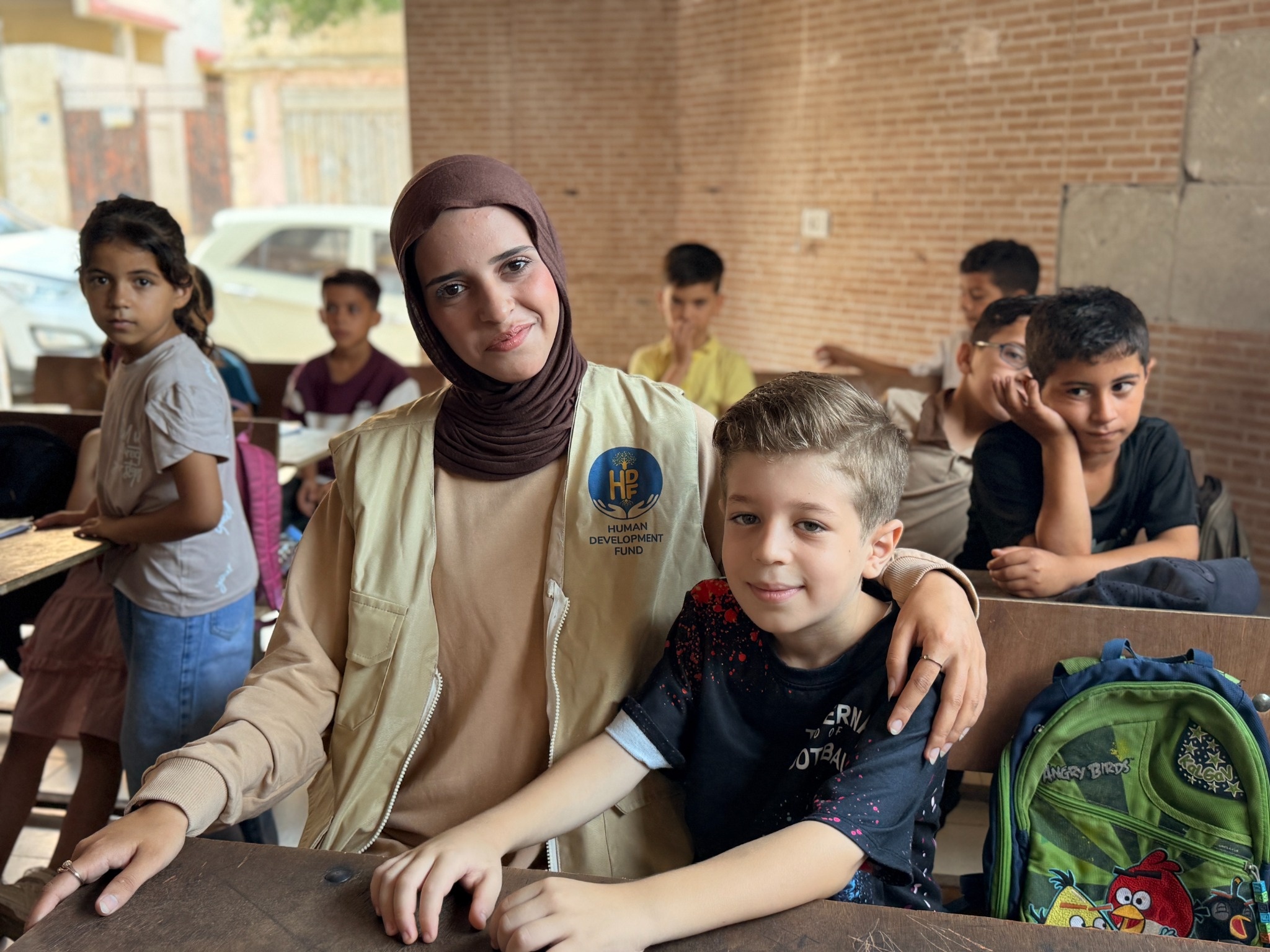 A woman sits with her arm around a boy at a classroom desk, with other children behind them.