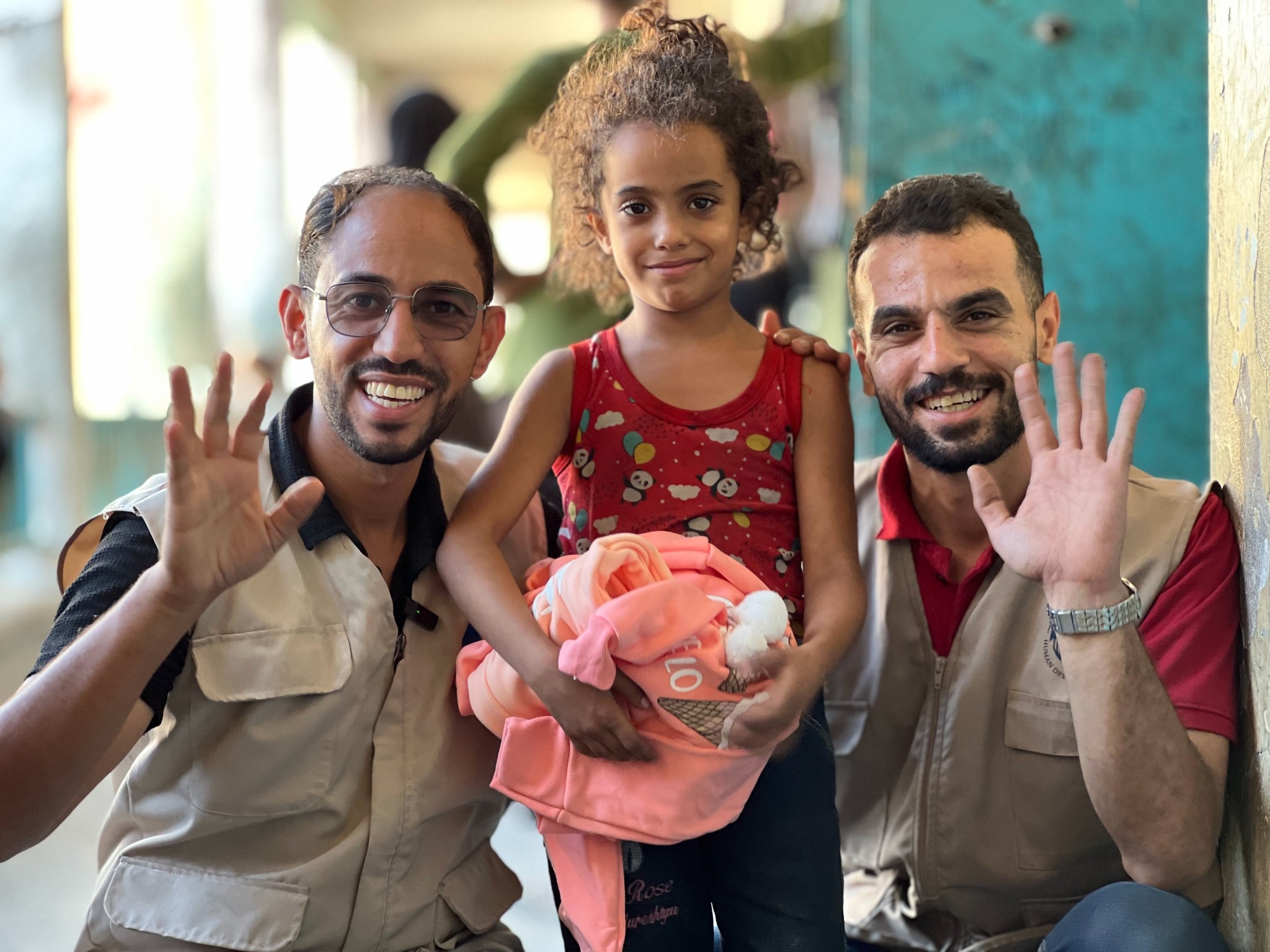 A girl holding pink clothing stands between two smiling men waving at the camera.