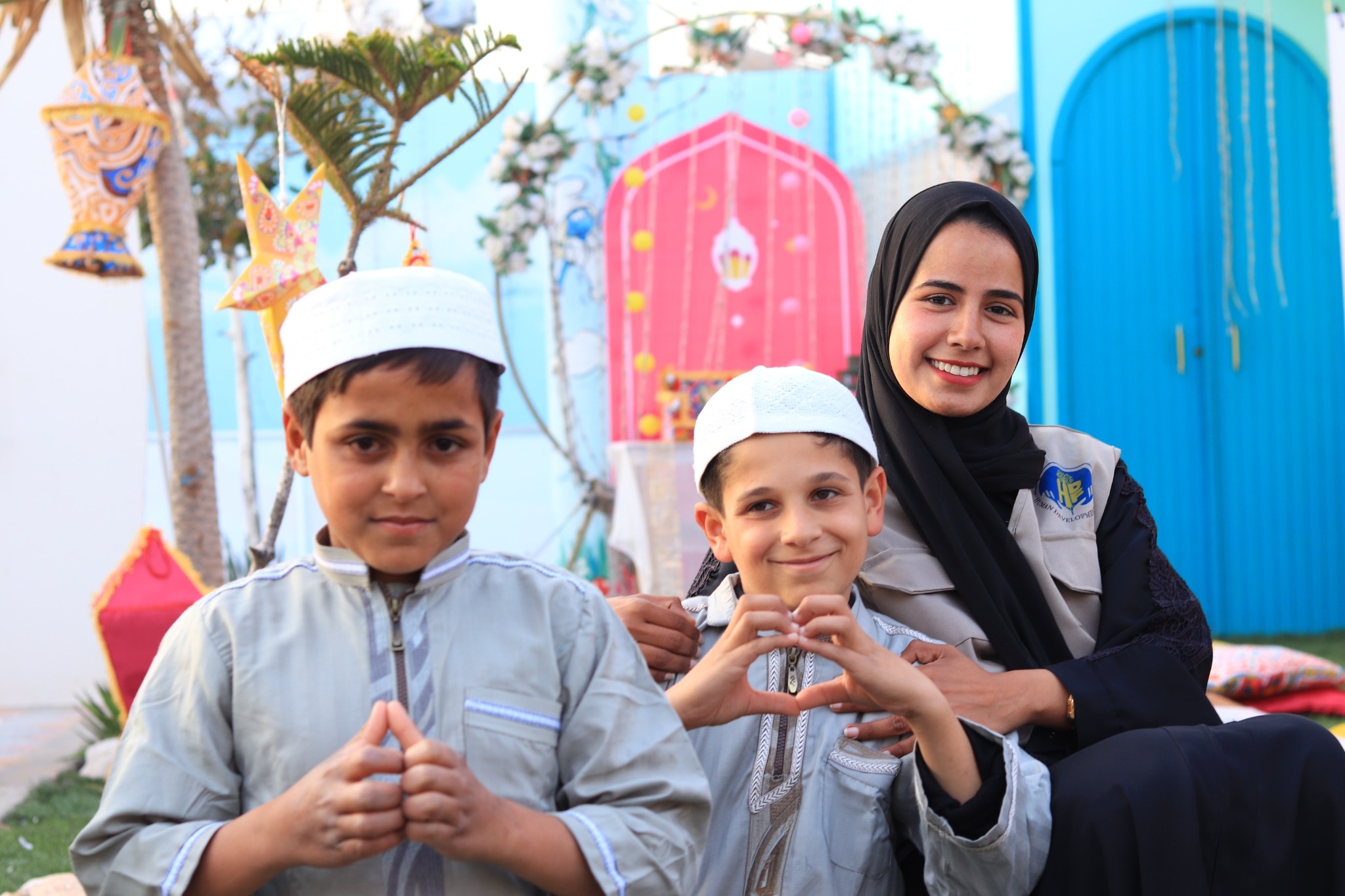 A woman poses with two boys in white caps against colorful decorations; one boy forms a heart with his hands.