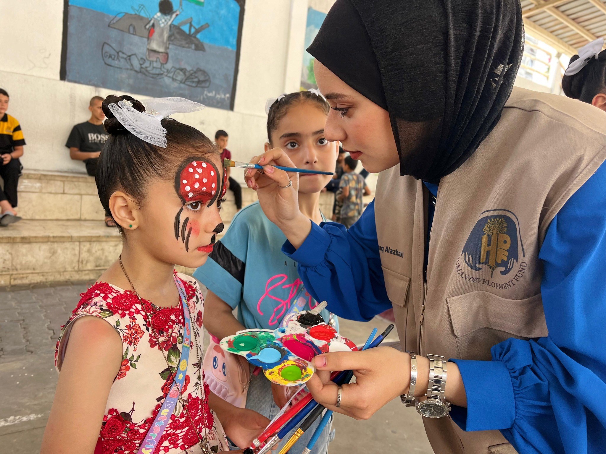 A woman paints a red, white, and black design on a girl’s face while another child watches.