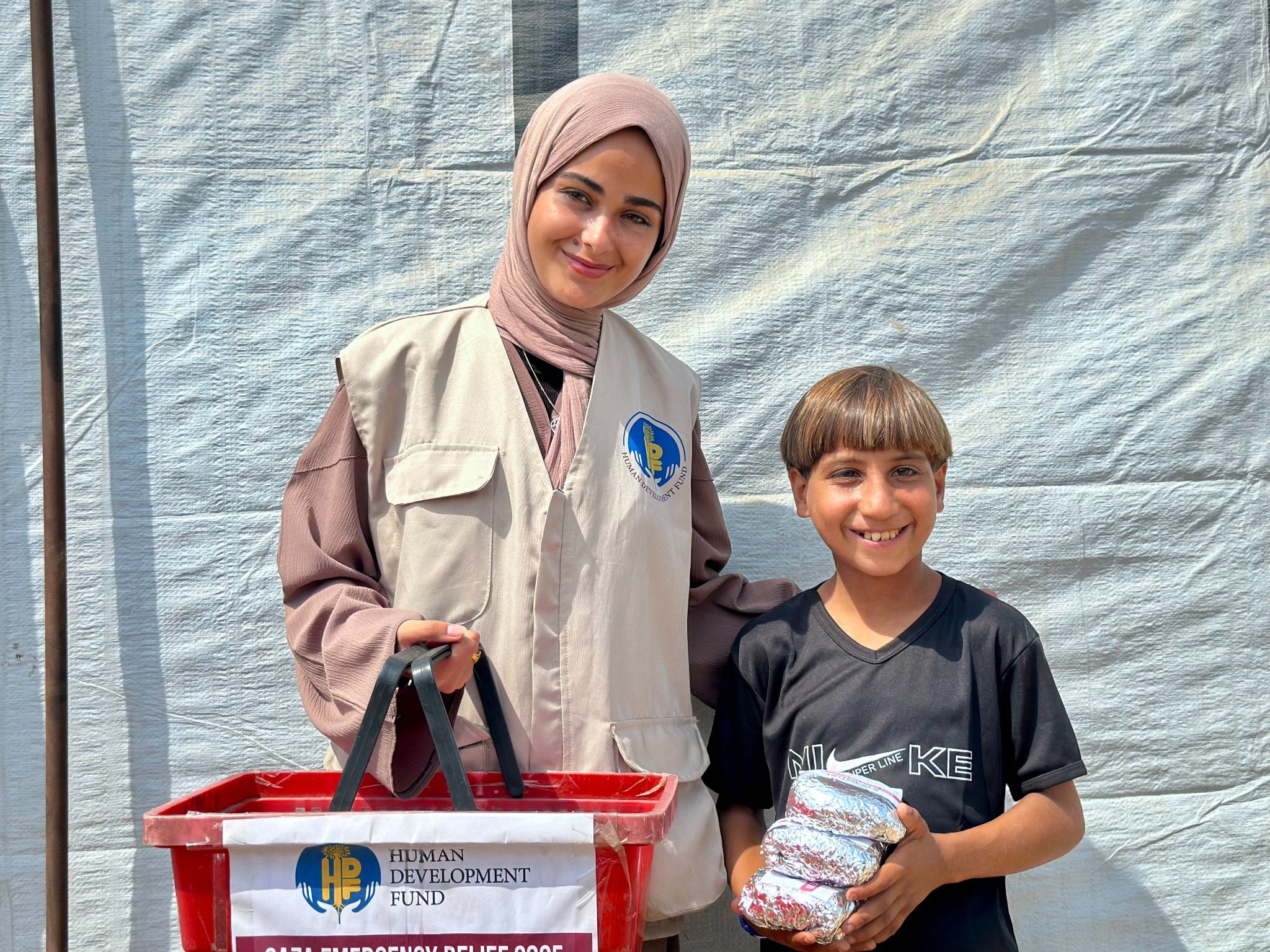 A woman holds a red basket beside a smiling boy carrying foil-wrapped meals in front of a tarp wall.